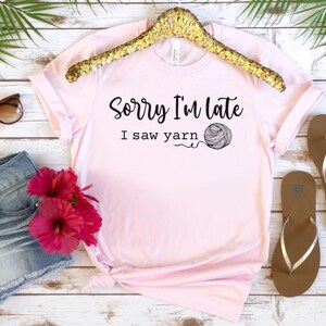 Op de afbeelding: Een roze T-shirt met de tekst "Sorry, ik ben te laat, ik zag garen" en een zwart-witte illustratie van een bol garen.