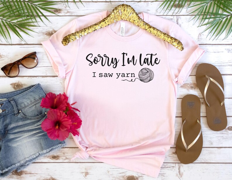 Op de afbeelding: Een roze T-shirt met de tekst "Sorry, ik ben te laat, ik zag garen" en een zwart-witte illustratie van een bol garen.