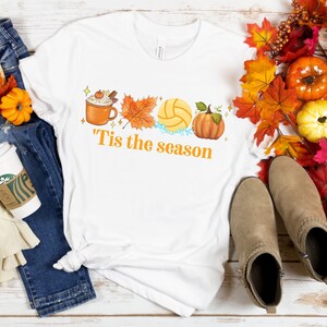 Pode incluir: Camiseta branca com um design gr&aacute;fico de uma x&iacute;cara de caf&eacute;, uma bola de v&ocirc;lei, uma ab&oacute;bora e folhas de outono. O texto "'Tis the season" est&aacute; impresso abaixo do gr&aacute;fico.