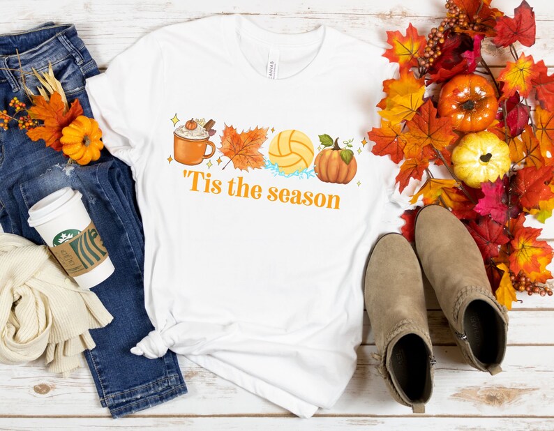 Pode incluir: Camiseta branca com um design gr&aacute;fico de uma x&iacute;cara de caf&eacute;, uma bola de v&ocirc;lei, uma ab&oacute;bora e folhas de outono. O texto "'Tis the season" est&aacute; impresso abaixo do gr&aacute;fico.