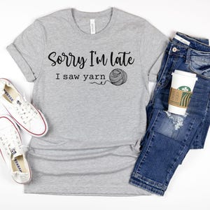 Op de afbeelding: Grijs T-shirt met de tekst "Sorry I'm late I saw yarn" en een afbeelding van een bol garen. Het shirt wordt gecombineerd met een blauwe jeans en witte sneakers.