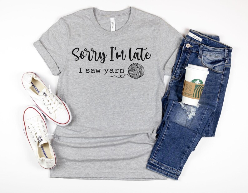 Op de afbeelding: Grijs T-shirt met de tekst "Sorry I'm late I saw yarn" en een afbeelding van een bol garen. Het shirt wordt gecombineerd met een blauwe jeans en witte sneakers.