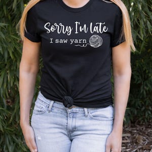 Op de afbeelding: Een zwart T-shirt met de tekst "Sorry, ik ben te laat, ik zag garen" en een witte omtrek van een bol garen.