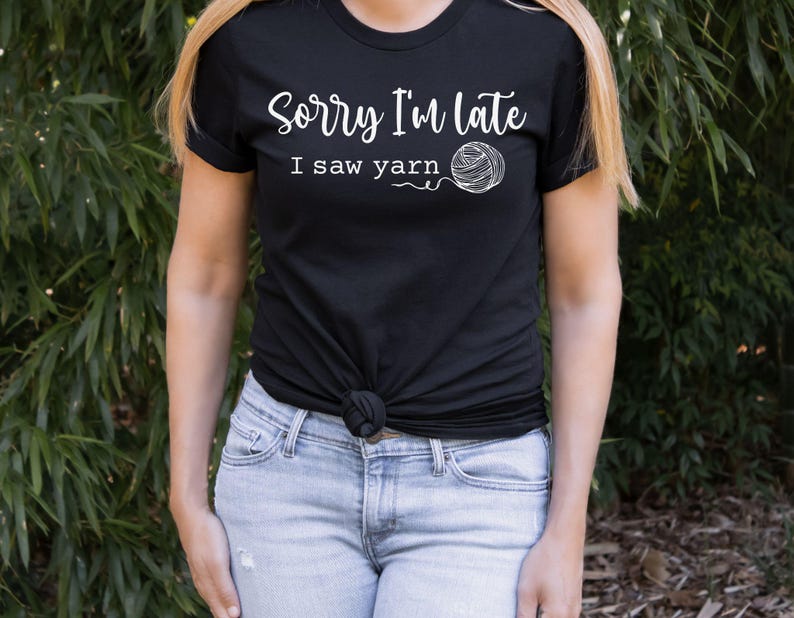 Op de afbeelding: Een zwart T-shirt met de tekst "Sorry, ik ben te laat, ik zag garen" en een witte omtrek van een bol garen.