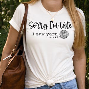 Op de afbeelding: Wit T-shirt met de tekst "Sorry I'm late I saw yarn" en een zwart-witte illustratie van een bol garen.