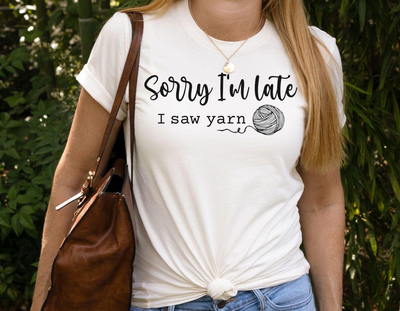 Op de afbeelding: Wit T-shirt met de tekst "Sorry I'm late I saw yarn" en een zwart-witte illustratie van een bol garen.