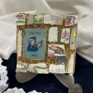 Puede incluir: Una obra de arte en mosaico con el tema del juego Monopoly. La pieza cuadrada está hecha de fragmentos de porcelana con diseños florales y estampados, purpurina dorada y una tarjeta de juego "GO TO JAIL". También se incluye una imagen de loro colorido.