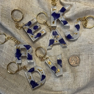 May include: A collection of resin letter keychains. Each letter is clear with blue and gold accents, attached to a gold-colored keyring. The letters are arranged on a textured, neutral-toned surface.