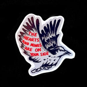 Könnte beinhalten: Ein Aufkleber mit einem Vogel mit ausgebreiteten Flügeln in Schwarz, Weiß und Rot. Der Text "THE HEARTS AND MINDS ARE ON YOUR SIDE" steht auf den Flügeln. Der Aufkleber hat einen weißen Rand.