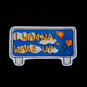 May include: A blue and white sign with the text "I wanna wake up" in yellow letters. The sign has a white border and is decorated with yellow flowers and white clouds.