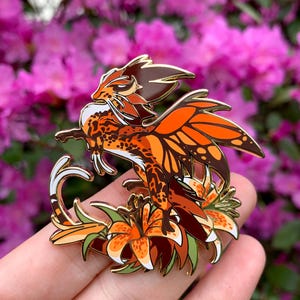 May include: A detailed enamel pin featuring a fantastical creature with orange and brown patterned wings and body, perched amidst orange lilies. The pin has gold-colored accents and is set against a blurred floral background.