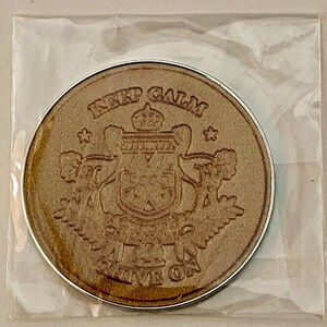 May include: A round, light brown coaster with a silver edge. The coaster features a crest design with the words "KEEP CALM" at the top and "CARRY ON" at the bottom. The coaster is in a clear plastic sleeve.