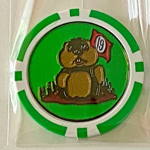 May include: A golf ball marker with a green and white border. The center features a cartoon groundhog with a red flag that has the number 19 on it. The groundhog is brown and tan, and the background is green.