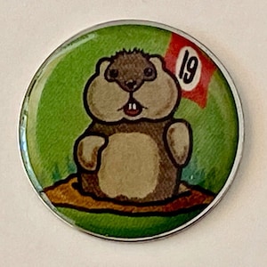 May include: A round, green and brown golf ball marker with a cartoon gopher and a red flag with the number 19 on it.