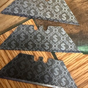 Könnte beinhalten: Drei silberne Cuttermesser-Klingen mit einem sich wiederholenden Dollarzeichenmuster. Die Klingen sind auf einer Holzoberfläche angeordnet. Die Klingen haben oben eine kleine Kerbe.