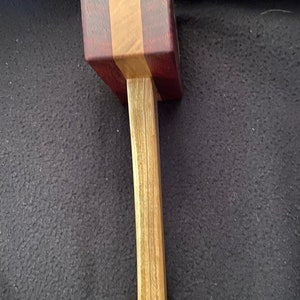 May include: A wooden mallet with a brown handle and a square head made of alternating strips of light and dark wood.