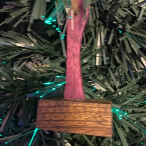 May include: A wooden Christmas ornament shaped like a hammer with a pink handle. The ornament is hanging on a green artificial Christmas tree with green and white lights.