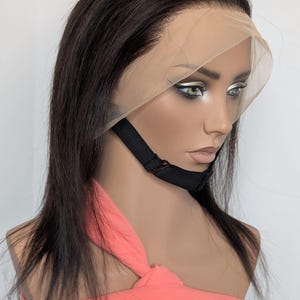 Peut inclure: Perruque brun foncé avec un devant en dentelle sur une tête de mannequin. La perruque présente de longs cheveux raides et une ligne de cheveux d'apparence naturelle. Une sangle noire maintient la perruque. Le mannequin porte un haut corail.