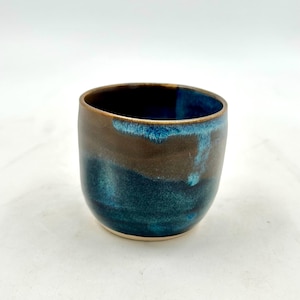 May include: A handmade ceramic cup with a rounded shape. The cup features a gradient of colors, transitioning from a dark brown rim to a deep teal blue at the base. The interior is a dark, contrasting color.