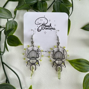 以下が含まれることがあります： 緑色のクリスタルアクセントが付いた甲虫のシルエットが特徴のシルバー製のイヤリングのペア。イヤリングは緑色のビーズの円に囲まれ、シルバーのフックからぶら下がっています。イヤリングは「Mush Creations」というテキストが書かれた白いカードに飾られています。