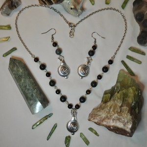 May include: A silver necklace with a snail pendant and matching earrings. The necklace features black beads and the earrings have green beads. The necklace and earrings are on a white surface with green crystals.