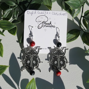 May include: A pair of silver ladybug earrings with black and white spots. Each earring features a black obsidian bead and a red dyed quartz bead hanging from it. The earrings are displayed on a white card with the text "Dyed quartz + Obsidian! Mush Creations".
