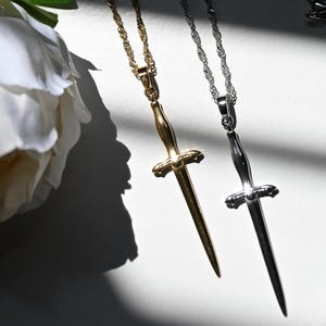 K&ouml;nnte beinhalten: Zwei Halsketten mit dolchf&ouml;rmigen Anh&auml;ngern. Eine ist goldfarben, die andere silberfarben, beide mit filigranen Kettengliedern. Im Hintergrund eine wei&szlig;e Blume, mit Schatten, die Tiefe verleihen.