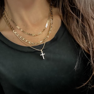 Puede incluir: Collares dorados en capas. La capa superior es una cadena delicada, la capa intermedia es una cadena más gruesa y la capa inferior presenta un colgante de cruz. Los collares se usan sobre una camisa negra de manga larga.