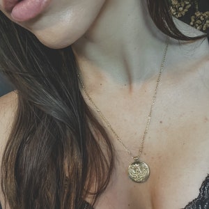 Gold chain necklace with a gold circular pendant. The pendant has an embossed design of Goddess Isis with wings spread wide. The necklace is shown against a dark background.