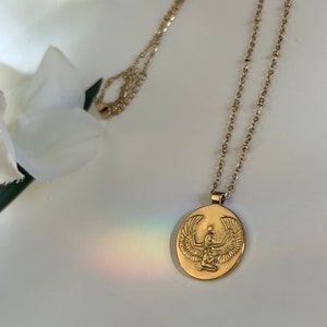 14k Gold necklace with an oval pendant featuring an embossed image of the Egyptian goddess Isis. The pendant is suspended from a delicate gold chain. The necklace is displayed against a white background, with a blurred image of white flowers in the upper left corner. The overall aesthetic is minimalist and elegant, suitable for layering or wearing alone.