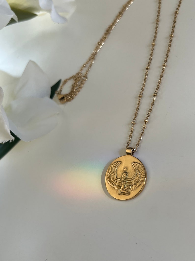 14k Gold necklace with an oval pendant featuring an embossed image of the Egyptian goddess Isis. The pendant is suspended from a delicate gold chain. The necklace is displayed against a white background, with a blurred image of white flowers in the upper left corner. The overall aesthetic is minimalist and elegant, suitable for layering or wearing alone.