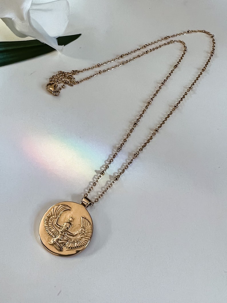 Gold necklace featuring a circular pendant with an embossed design of Isis, the Egyptian goddess with outstretched wings. The chain is delicate and features small beads. The pendant is a rich gold tone, and the overall style is elegant and sophisticated.  This gold statement necklace is perfect for adding a touch of ancient Egyptian mythology to any outfit.