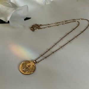 K&ouml;nnte beinhalten: Goldfarbene Halskette mit einem kreisf&ouml;rmigen Anh&auml;nger mit einem gepr&auml;gten Vogeldesign. Die Kette ist filigran und der Anh&auml;nger ist mittig. Die Halskette wird auf einer wei&szlig;en Oberfl&auml;che mit einer wei&szlig;en Blume im Hintergrund pr&auml;sentiert.