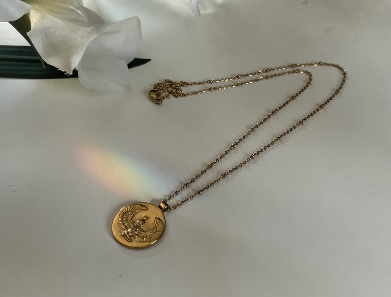 K&ouml;nnte beinhalten: Goldfarbene Halskette mit einem kreisf&ouml;rmigen Anh&auml;nger mit einem gepr&auml;gten Vogeldesign. Die Kette ist filigran und der Anh&auml;nger ist mittig. Die Halskette wird auf einer wei&szlig;en Oberfl&auml;che mit einer wei&szlig;en Blume im Hintergrund pr&auml;sentiert.