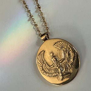Gold necklace with an oval pendant featuring an embossed design of a winged Isis figure. The chain is delicate and features small beads. The pendant is detailed and shows intricate craftsmanship.  The necklace is a gold tone metal and the pendant is a polished finish.  The style is elegant and mystical.
