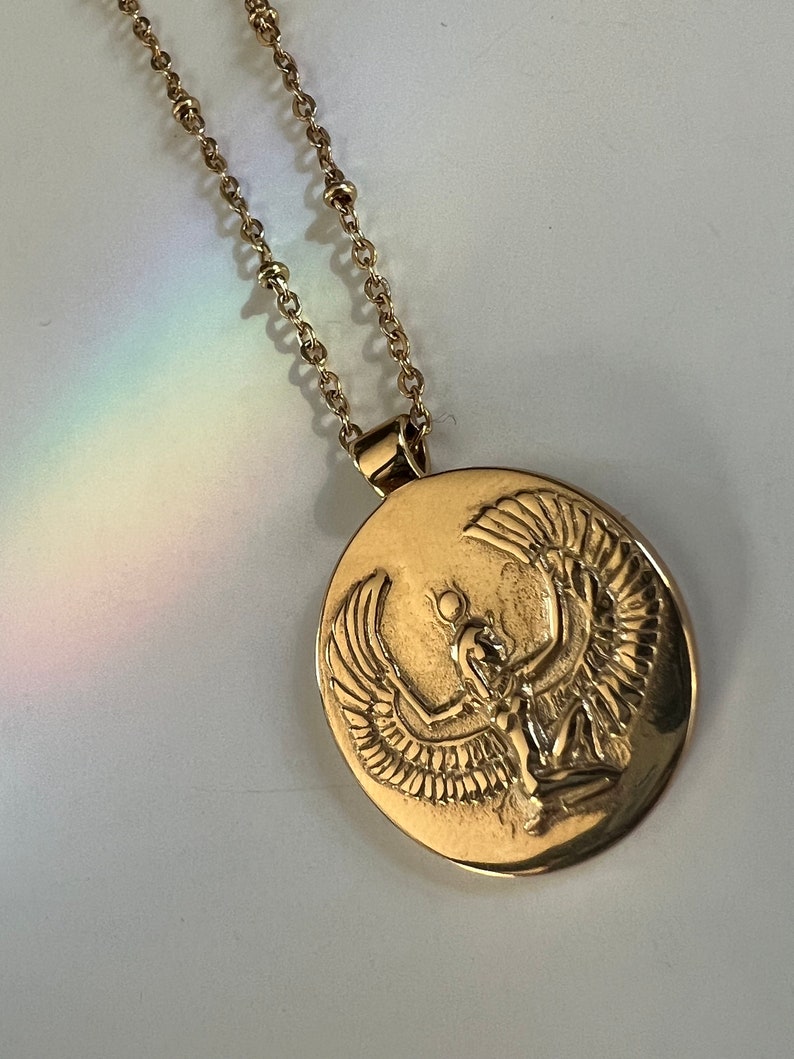 Gold necklace with an oval pendant featuring an embossed design of a winged Isis figure. The chain is delicate and features small beads. The pendant is detailed and shows intricate craftsmanship.  The necklace is a gold tone metal and the pendant is a polished finish.  The style is elegant and mystical.