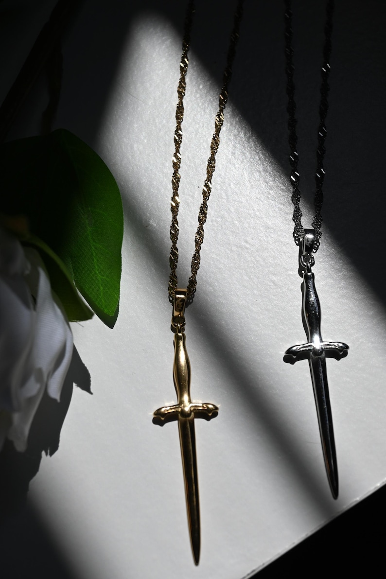K&ouml;nnte beinhalten: Zwei Halsketten mit Anh&auml;ngern in Dolchform. Eine ist goldfarben, die andere silberfarben, beide h&auml;ngen an filigranen Ketten. Das Bild ist vor einem wei&szlig;en Hintergrund mit Schatten platziert. Eine wei&szlig;e Blume und ein gr&uuml;nes Blatt sind im Hintergrund.