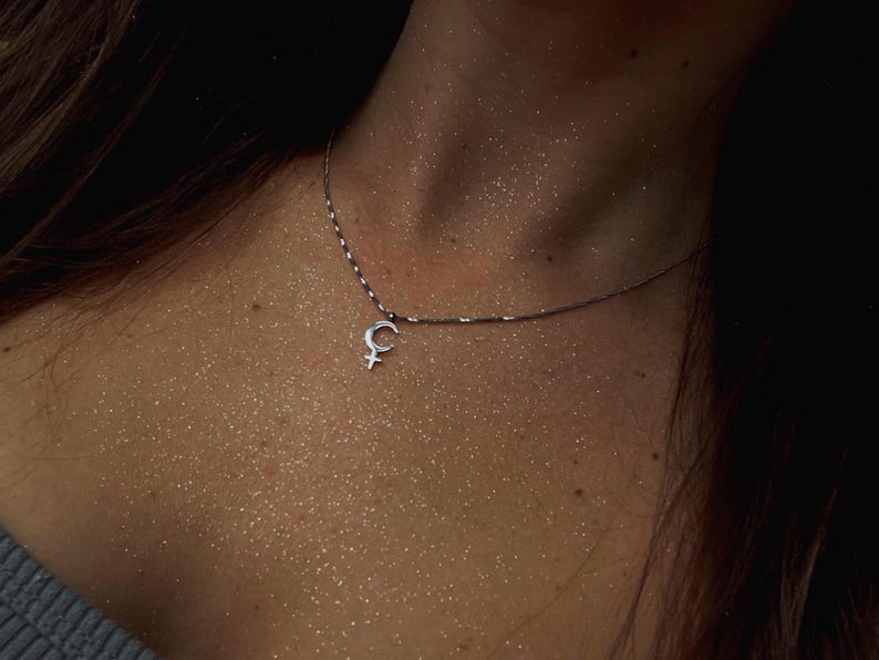May include: A silver necklace featuring a pendant with the female symbol. The pendant is a crescent moon shape with a cross below it. The necklace is worn on a person's neck.