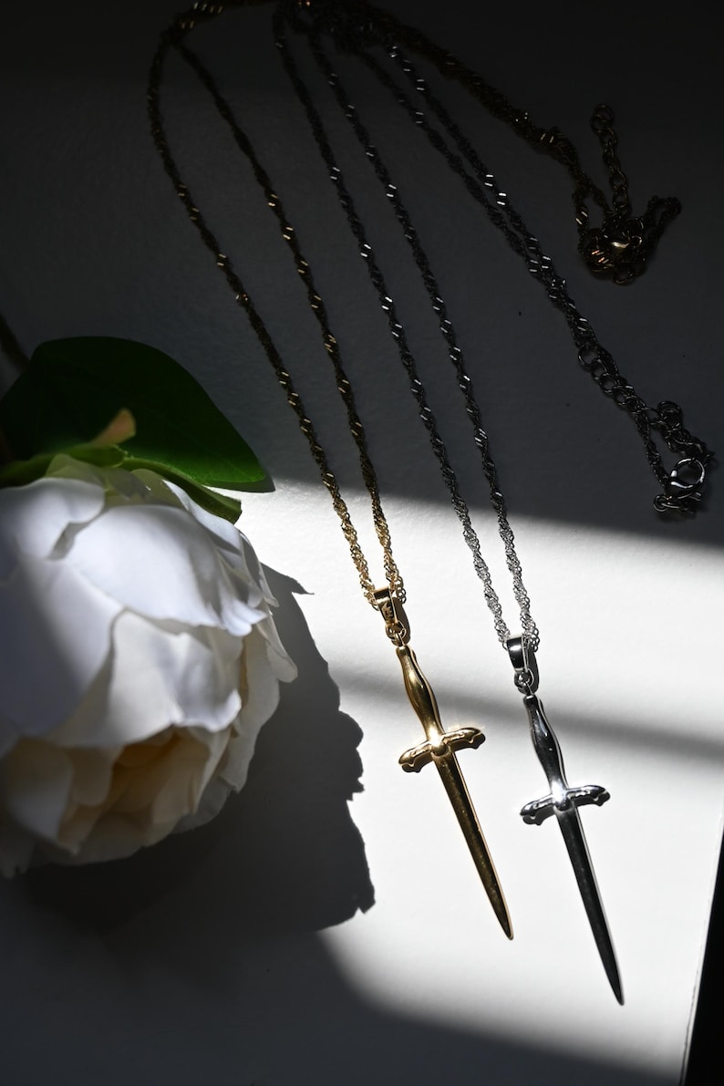 K&ouml;nnte beinhalten: Zwei Halsketten mit dolchf&ouml;rmigen Anh&auml;ngern. Eine ist goldfarben, die andere silberfarben, beide mit filigranen Kettengliedern. Im Hintergrund eine wei&szlig;e Blume, mit Schatten, die Tiefe verleihen.