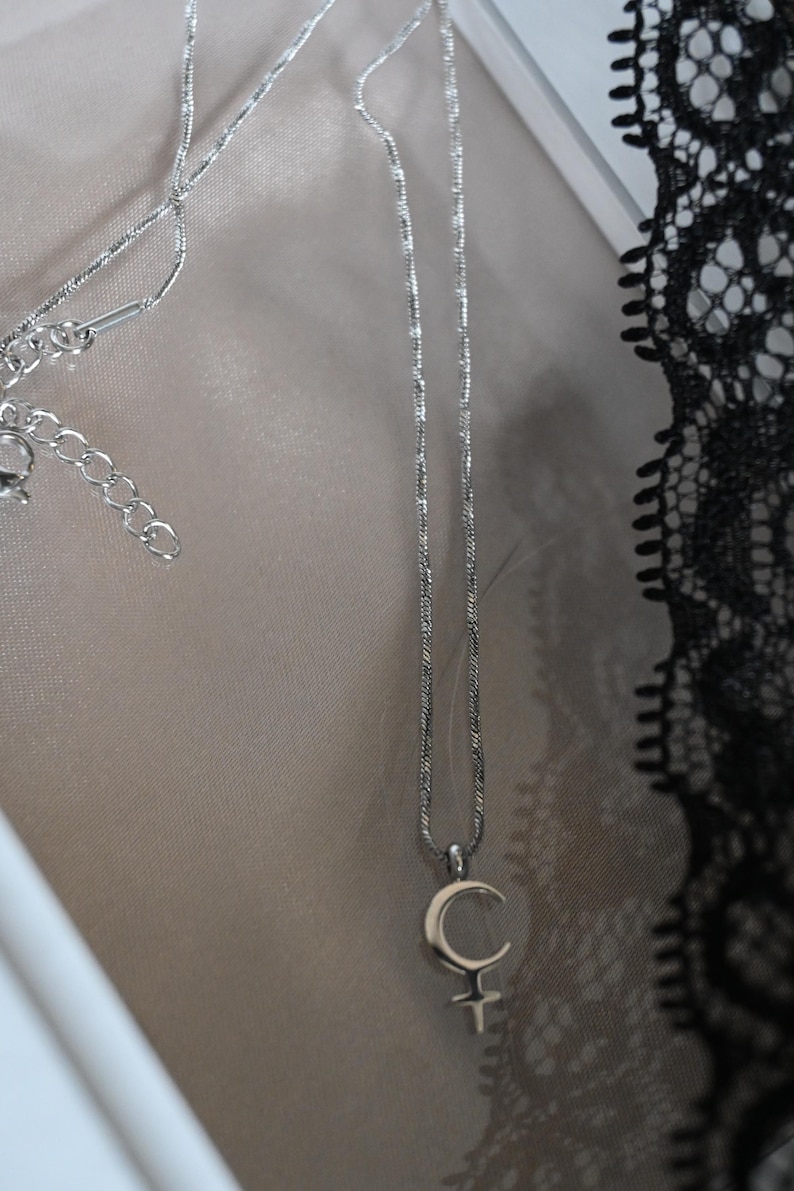 May include: A silver-toned necklace with a crescent moon and cross pendant. The chain is delicate and hangs against a neutral background. The necklace is a piece of jewelry.