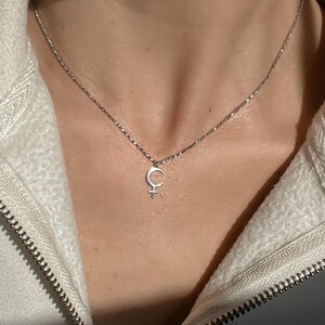 May include: Silver-toned necklace featuring a crescent moon and star pendant. The delicate chain is visible against a white jacket. The pendant is a symbol of femininity. The necklace is a piece of jewelry.