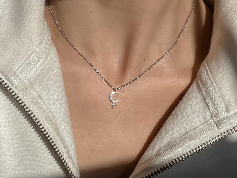 May include: Silver-toned necklace featuring a crescent moon and star pendant. The delicate chain is visible against a white jacket. The pendant is a symbol of femininity. The necklace is a piece of jewelry.