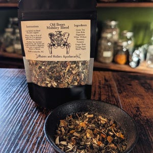 May include: A black pouch of "Old Bones Mobility Blend" herbal tea, with a black bowl of tea next to it. The label lists ingredients like green tea and turmeric. The background shows shelves with jars.