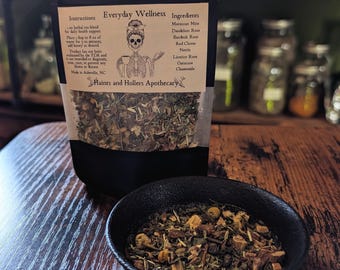 May include: A black pouch of "Everyday Wellness" herbal tea blend with a skull illustration, and a small black bowl filled with loose tea. The ingredients listed are Moroccan Mint, Dandelion Root, Burdock Root, Red Clover, Nettle, Licorice Root, Oatstraw, and Chamomile.