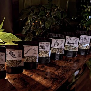 May include: A collection of sealed black pouches filled with dried herbs and labeled with white text and illustrations. The pouches are arranged on a dark wooden surface, with green plants in the background. The labels include text such as "Red Moon Tea" and "Soothing Herbal Tea".