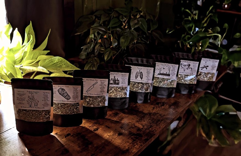 May include: A collection of sealed black pouches filled with dried herbs and labeled with white text and illustrations. The pouches are arranged on a dark wooden surface, with green plants in the background. The labels include text such as "Red Moon Tea" and "Soothing Herbal Tea".