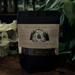 May include: A black stand-up pouch containing loose-leaf tea, labeled Haints and Hollers Apothecary Whispers from the Gods Below. The label features a cave illustration, brewing instructions for 8 oz of water, and ingredients including black tea, chicory, vanilla rooibos, cacao nibs, espresso beans, cinnamon, and cardamom.