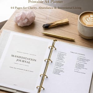 May include: An open Manifestation Journal with gold binder rings, a gold pen, and a cup of coffee with latte art. A pink crystal and a piece of Palo Santo are also on the wooden surface. The journal is a printable A4 planner.