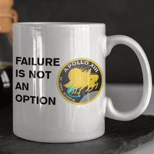 Könnte beinhalten: Eine weiße Keramik-Kaffeetasse mit einer schwarzen Aufschrift, die "Scheitern ist keine Option" lautet. Die Tasse zeigt außerdem einen gelben und schwarzen Apollo-13-Missions-Patch mit dem Text "Apollo XIII Ex Luna Scientia".