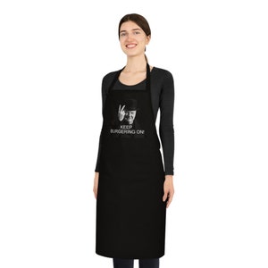 May include: Black apron featuring a black and white image of a man in a hat giving the victory sign, with the text "KEEP BURGERING ON!". The apron is worn by a person.